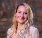 Smiling blonde woman with long hair wearing a light-colored top against a blurred brownish background.