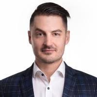 Headshot of a man with short brown hair wearing a white shirt and dark checkered blazer against a white background.