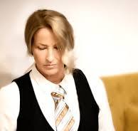 Blonde woman wearing a striped tie, white shirt, and black vest looking downward against a blurred background.