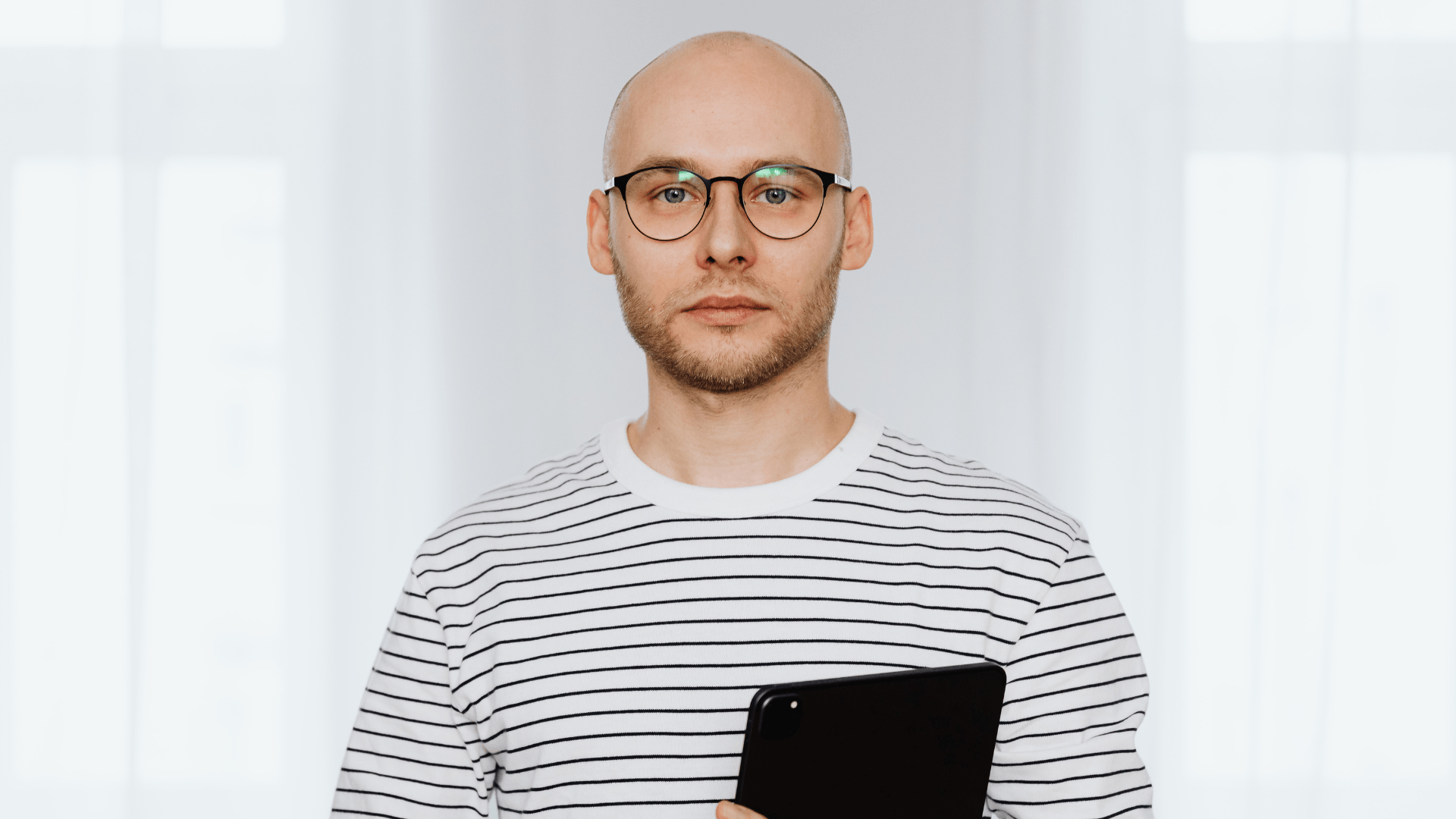 Bald man with glasses wearing a white and black striped shirt holding a black tablet.