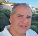 Middle-aged man with short gray hair smiling outdoors with greenery and buildings in the background.