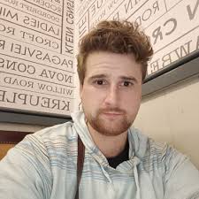 Young man with light brown hair and beard wearing a light-colored hoodie sitting indoors with framed text art on the walls behind him.